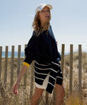 A woman in a navy Cashmere Marina Crewneck, white cap, and striped sweater tied at her waist stands by a wooden fence, with dry grass, blue sky, and the sea in the background.