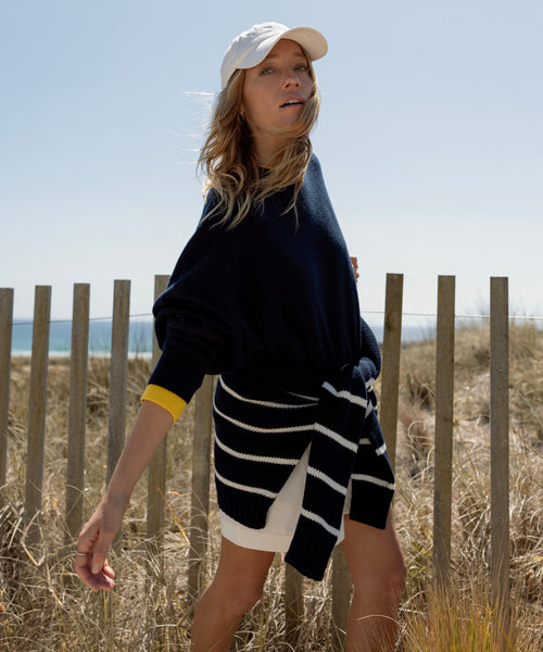 A woman in a navy Cashmere Marina Crewneck, white cap, and striped sweater tied at her waist stands by a wooden fence, with dry grass, blue sky, and the sea in the background.