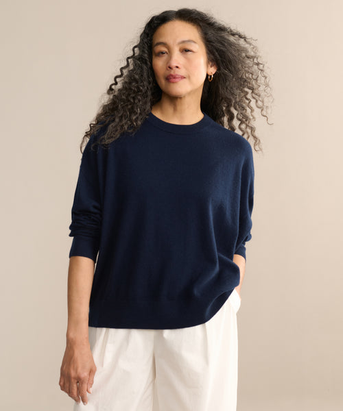 A woman with long curly hair wears the Cashmere Marina Crewneck in navy blue and white pants, standing against a beige background and looking confidently at the camera with one hand in her pocket.