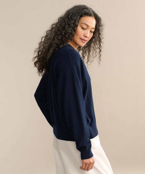 A woman with long, curly hair stands sideways against a beige background, wearing the Cashmere Marina Crewneck in navy blue and white pants. She looks down with a calm expression.