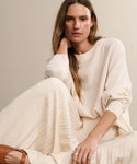 A woman with long, light brown hair sits against a beige background, wearing the oversized Cashmere Marina Crewneck and a pleated cream skirt. She looks relaxed, resting her head on her hand and embodying effortless style.