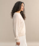 A woman with long, curly hair stands in profile against a beige background, wearing the Cashmere Marina Crewneck in light beige paired with a white skirt.