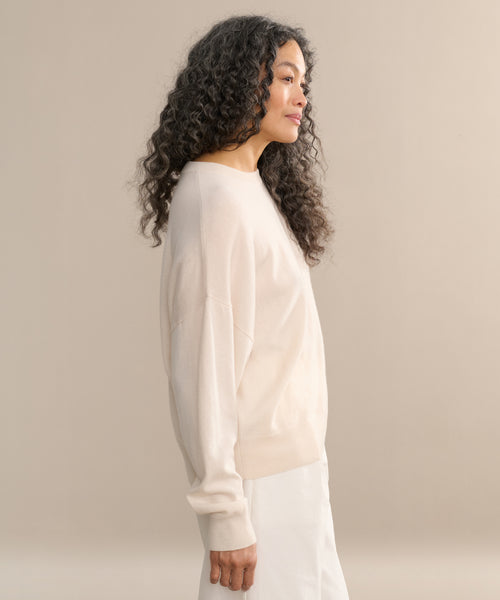 A woman with long, curly hair stands in profile against a beige background, wearing the Cashmere Marina Crewneck in light beige paired with a white skirt.
