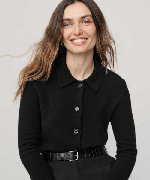 A woman with long brown hair smiles at the camera, wearing the Murphy Cardigan and black pants with a black belt, standing against a light gray background.