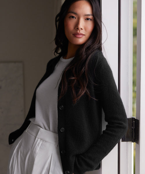 A woman with long dark hair confidently leans against a door frame, wearing the Murphy Cardigan in black over a white shirt and high-waisted white pants.