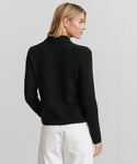 A woman with blonde hair models the Murphy Cardigan, a black knit sweater, paired with white pants. She stands against a plain light background, facing away with her head turned slightly to the side.