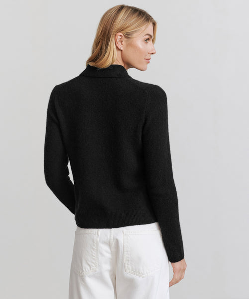 A woman with blonde hair models the Murphy Cardigan, a black knit sweater, paired with white pants. She stands against a plain light background, facing away with her head turned slightly to the side.