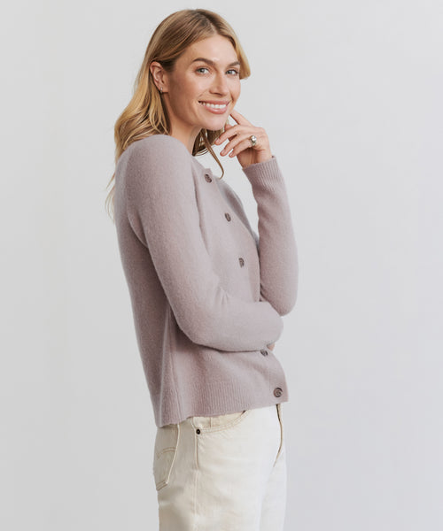 A smiling blonde woman poses against a light background, wearing the Murphy Cardigan in light purple and cream pants, with her hand near her chin.
