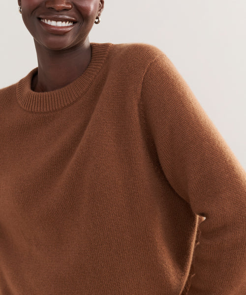 A person is smiling, wearing the Cashmere Oversized Crewneck in soft brown with a relaxed fit. Only their upper body and part of their face are visible against a plain, light background.