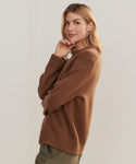 A woman with blonde hair stands sideways against a plain light background, softly smiling and touching her chin while wearing the Cashmere Oversized Crewneck sweater and olive pants.