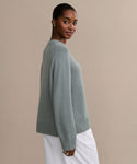 A woman with pulled-back hair models the Cashmere Oversized Crewneck in light gray-green, paired with white pants. She stands sideways, looking at the camera against a plain beige background.