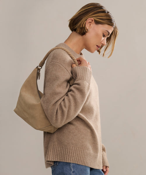 A woman with short, light brown hair wears the Cashmere Oversized Crewneck in beige and blue jeans, carrying a tan shoulder bag. She is looking down while standing against a plain, light-colored background.