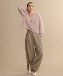 A woman poses against a beige background, wearing the Cashmere Peyton Cardigan in soft pink with puff sleeves and large buttons, paired with loose taupe wide-leg trousers and white shoes. One arm is bent, her hand resting on her head.
