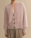 A person wears the Cashmere Peyton Cardigan in soft, light pink with a deep V-neck and long sleeves, styled with high-waisted beige pleated pants against a neutral background.
