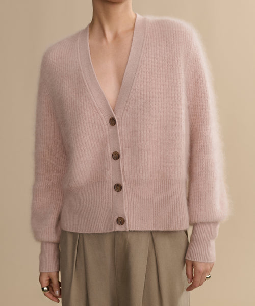 A person wears the Cashmere Peyton Cardigan in soft, light pink with a deep V-neck and long sleeves, styled with high-waisted beige pleated pants against a neutral background.