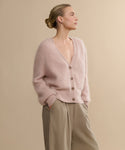 A blonde woman with a low bun wears the Cashmere Peyton Cardigan in soft pink and beige trousers, standing with hands in pockets against a plain beige background, gazing to the side.