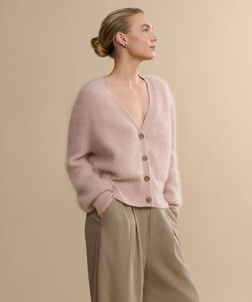 A blonde woman with a low bun wears the Cashmere Peyton Cardigan in soft pink and beige trousers, standing with hands in pockets against a plain beige background, gazing to the side.
