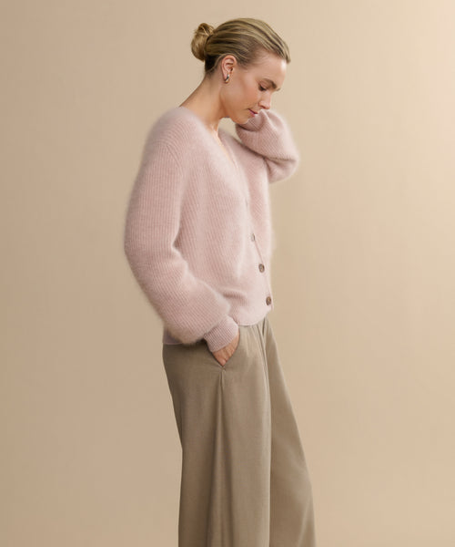 A woman in profile stands against a beige background, wearing the Cashmere Peyton Cardigan in soft pink with puff sleeves and light brown wide-leg pants. Her hair is styled in a neat bun as she looks down thoughtfully, one hand tucked into her pocket.