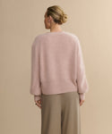 A person with blonde hair in a low bun is shown from the back, wearing a Cashmere Peyton Cardigan in soft pink with long sleeves and loose beige trousers, standing against a plain beige background.