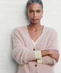 A woman with gray hair pulled back stands confidently with arms crossed, wearing a pale pink Cashmere Peyton Cardigan over a cream top against a light background.