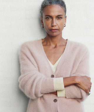 A woman with gray hair pulled back stands confidently with arms crossed, wearing a pale pink Cashmere Peyton Cardigan over a cream top against a light background.