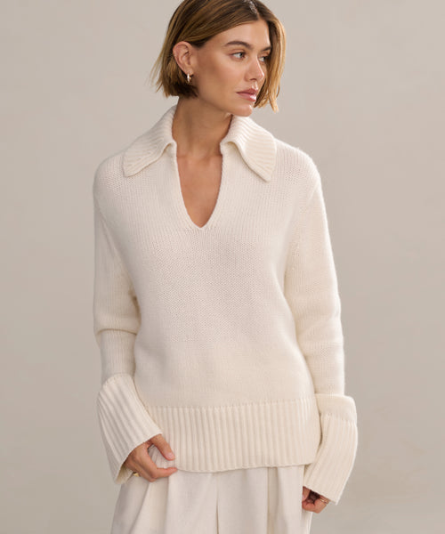 A woman with short light brown hair wears the Cashmere Phoebe Polo, an ivory sweater with a wide collar and ribbed cuffs, paired with a matching cream skirt, standing against a plain neutral background.