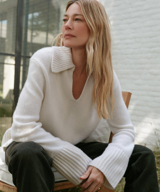 A woman with long blonde hair sits outdoors on a wooden chair, wearing the Cashmere Phoebe Polo with wide sleeves and dark pants. She gazes thoughtfully into the distance, framed by a white brick wall and glass window in the background.