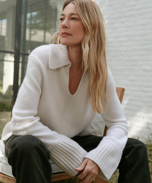 A woman with long blonde hair sits outdoors on a wooden chair, wearing the Cashmere Phoebe Polo with wide sleeves and dark pants. She gazes thoughtfully into the distance, framed by a white brick wall and glass window in the background.