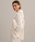A woman with short, light brown hair stands sideways, looking at the camera. She wears the Cashmere Phoebe Polo in ivory with matching pants and carries an off-white shoulder bag against a plain, neutral background.