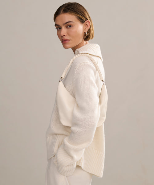 A woman with short, light brown hair stands sideways, looking at the camera. She wears the Cashmere Phoebe Polo in ivory with matching pants and carries an off-white shoulder bag against a plain, neutral background.