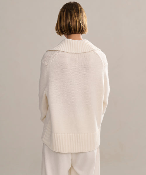 A person with short, light brown hair is seen from behind wearing the Cashmere Phoebe Polo, an ivory, loose-fit sweater with a wide collar and ribbed hem, paired with matching cream pants against a plain background.