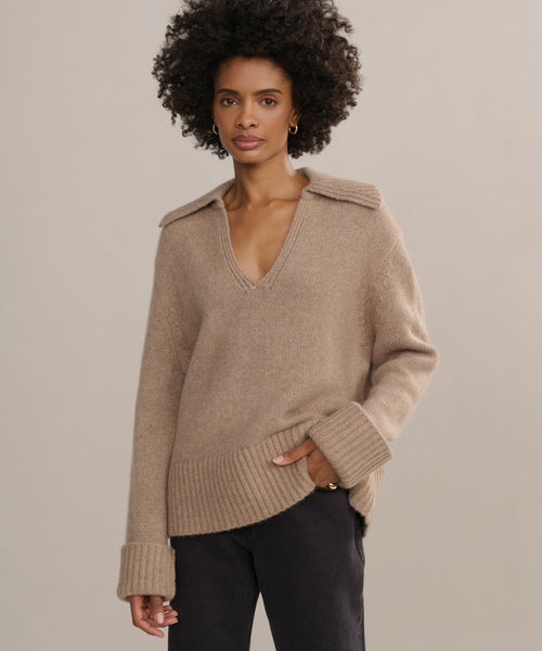 A woman with short curly hair wears the loose, tan Cashmere Phoebe Polo featuring a wide collar and rolled-up sleeves, paired with dark pants, while standing against a neutral background.