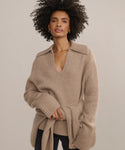 A woman with curly hair wears the beige Cashmere Phoebe Polo with a wide collar and long sleeves, standing confidently against a plain light background.