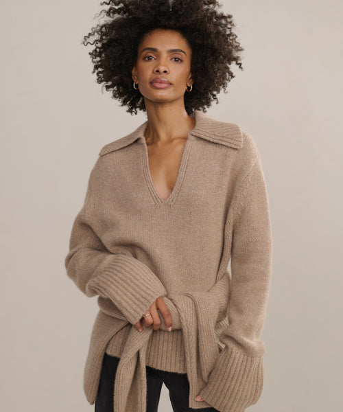 A woman with curly hair wears the beige Cashmere Phoebe Polo with a wide collar and long sleeves, standing confidently against a plain light background.