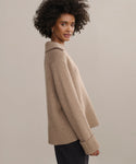 A woman with curly hair wears the Cashmere Phoebe Polo, a loose, tan cashmere sweater with a wide collar and ribbed cuffs and hem. She stands in profile against a light background, looking over her shoulder and smiling softly.