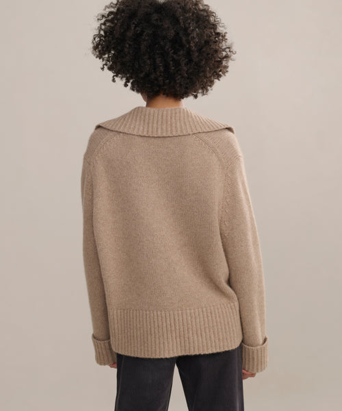 A person with curly hair, seen from behind, is wearing the beige Cashmere Phoebe Polo sweater with a wide collar and ribbed cuffs, paired with dark pants against a plain neutral background.