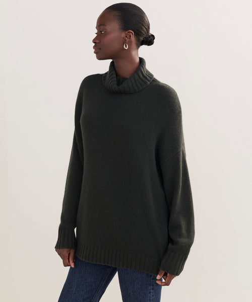 Wearing the dark oversized Cashmere Porter Turtleneck and blue jeans, a person with pulled-back hair and hoop earrings stands facing left against a plain light background.