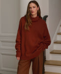 A woman with long brown hair stands indoors by a staircase, wearing the Cashmere Porter Turtleneck and wide-legged brown pants. She looks calmly at the camera, one hand in her pocket, effortlessly stylish.