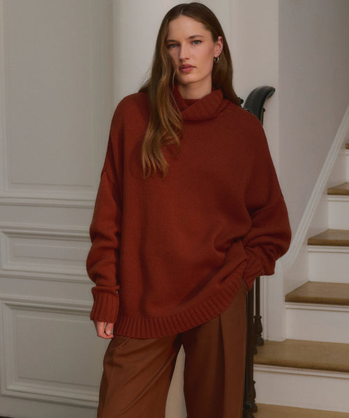 A woman with long brown hair stands indoors by a staircase, wearing the Cashmere Porter Turtleneck and wide-legged brown pants. She looks calmly at the camera, one hand in her pocket, effortlessly stylish.