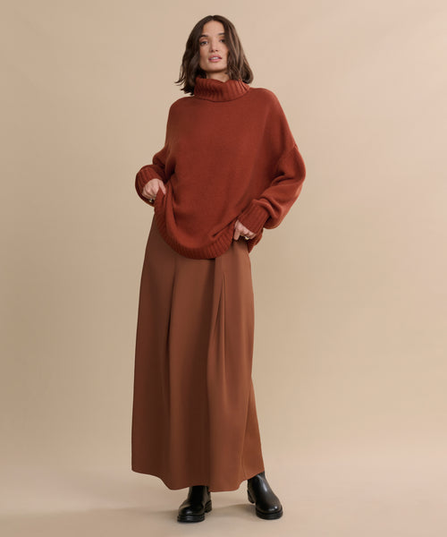 A person stands against a beige background, wearing the oversized Cashmere Porter Turtleneck with wide-legged brown pants and black boots, posing in a relaxed and confident manner.
