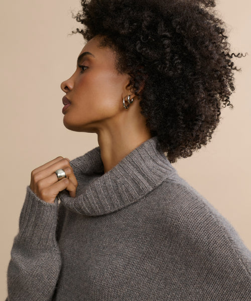 A woman with curly hair wears the oversized Cashmere Porter Turtleneck, paired with silver hoop earrings and a silver ring. Shown in profile against a beige background, she gently holds the sweater’s collar near her neck.