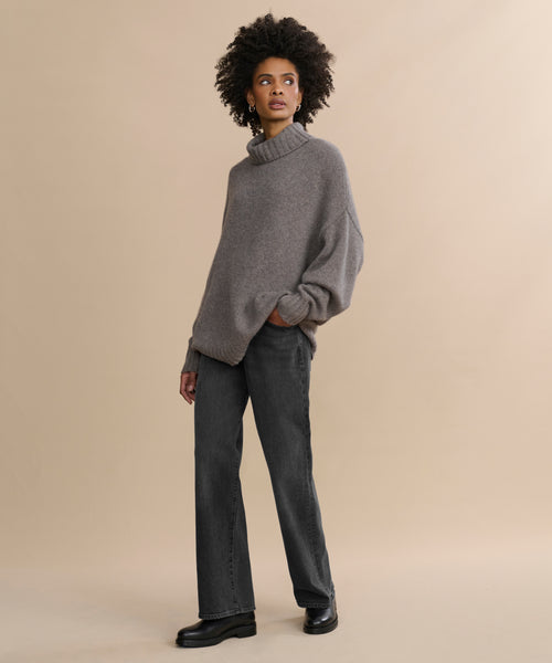 A person stands against a beige background wearing the Cashmere Porter Turtleneck, paired with dark jeans and black shoes. One hand is in their pocket as they look off to the side with a neutral expression.