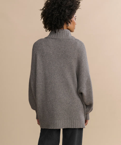 A person with curly hair is seen from the back, wearing the Cashmere Porter Turtleneck—an oversized, loose turtleneck sweater—paired with dark pants, standing against a plain beige background.