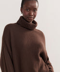 A woman with dark skin and pulled-back hair models the Cashmere Porter Turtleneck, an oversized sweater, while standing confidently against a plain light background.