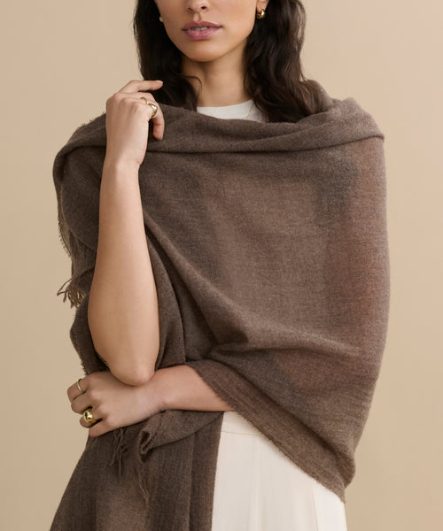 A woman drapes a Cashmere Shawl over her shoulders, paired with a cream outfit and gold rings against a beige backdrop. Her face is partly visible—an elegant, travel-ready look.