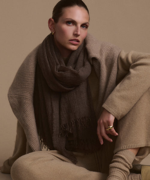 A person with slicked-back hair sits cross-legged against a beige background, wearing neutral-toned clothes with a long beige sweater, matching pants, gold hoop earrings, a ring, and a large dark brown Cashmere Shawl.