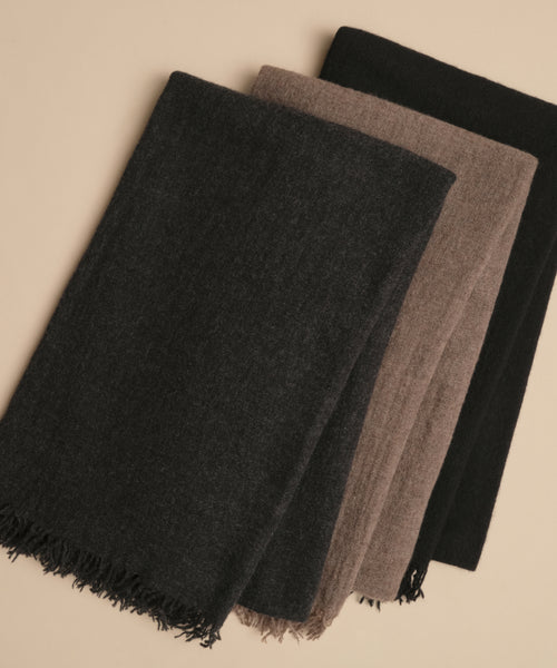 Three folded Cashmere Shawls in dark grey, brown, and black are laid side by side on a beige surface. The left shawl has fringed edges, while the others have plain ends—each one a perfect lightweight travel accessory.