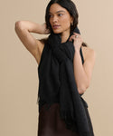 A woman with long dark hair wears a sleeveless black top and a lightweight Cashmere Shawl, draped and knotted around her neck as she poses with one arm bent behind her head against a beige background.