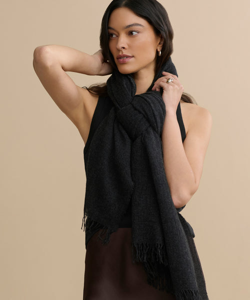 A woman with long dark hair wears a sleeveless black top and a lightweight Cashmere Shawl, draped and knotted around her neck as she poses with one arm bent behind her head against a beige background.