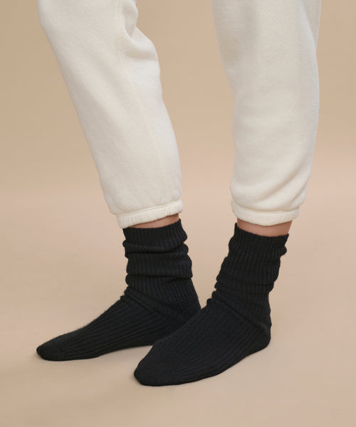 A person wears off-white sweatpants with ankles slightly rolled up, highlighting luxurious Cashmere Socks in black. Standing on a neutral beige background, the socks are perfectly showcased—making them an ideal gift choice.